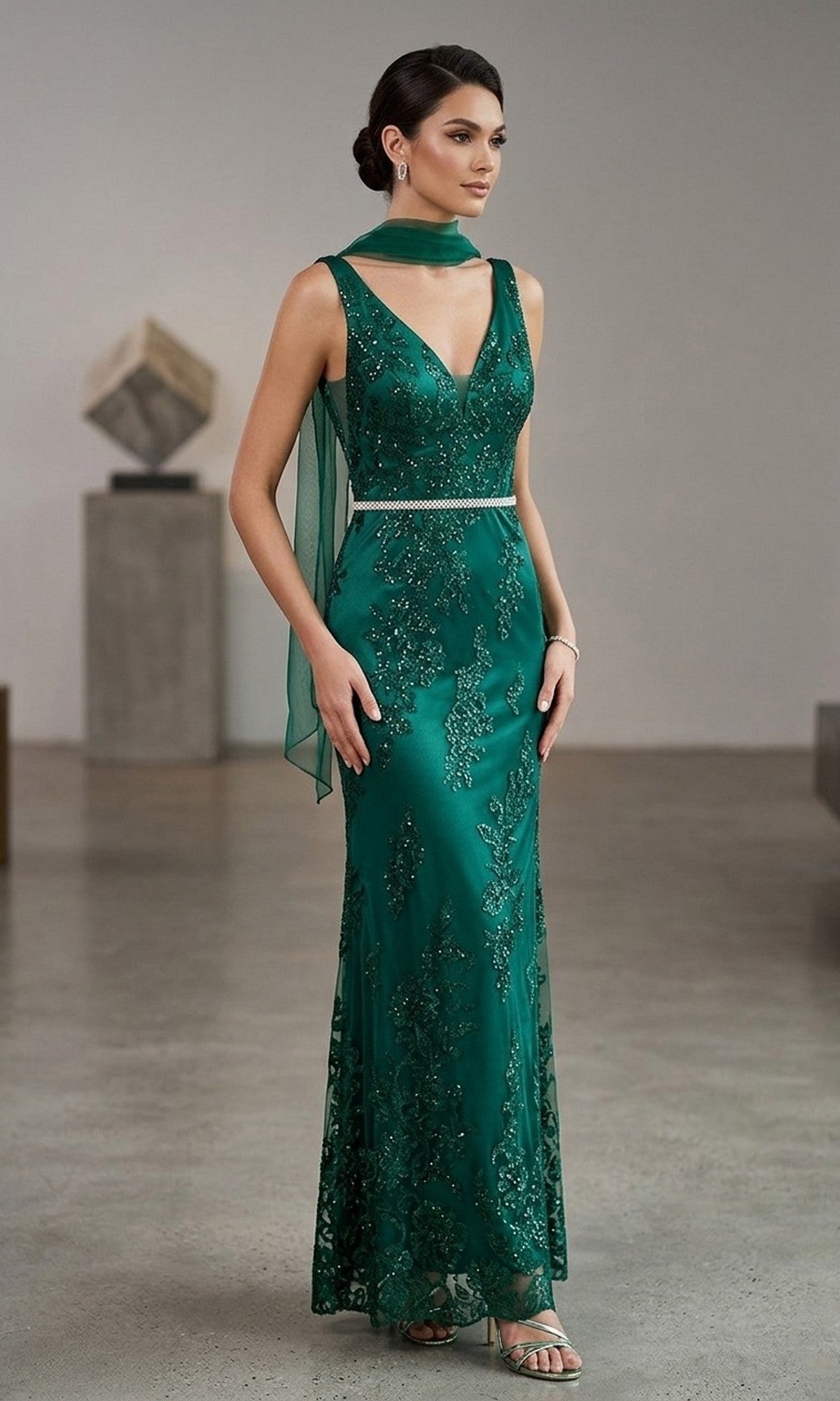 A woman stands indoors wearing the Chicas DM4026 long formal dress in emerald green, adorned with sparkling embellishments and an embroidered tulle overlay. She completes her look with a matching scarf, a silver belt, and strappy heels, with her hair elegantly styled in an updo.